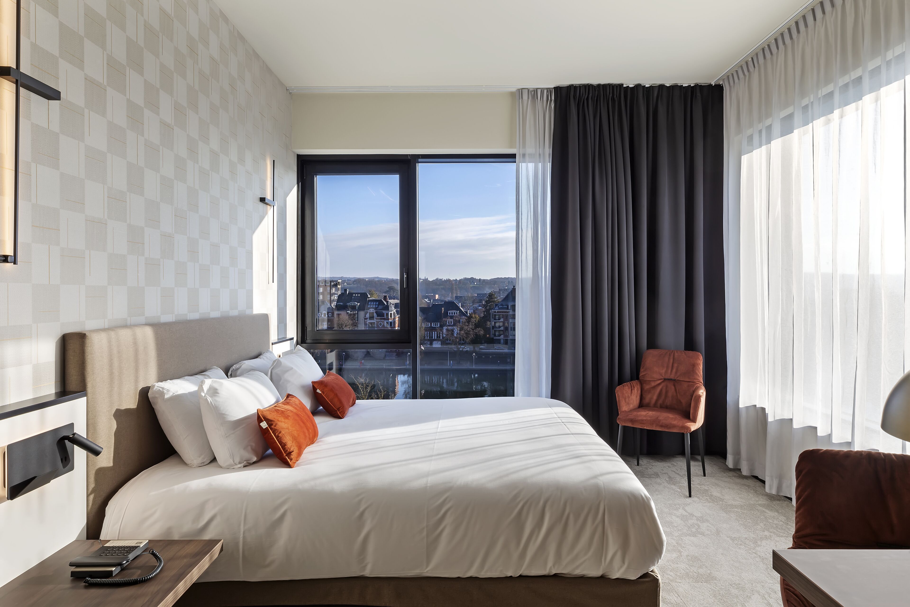 Habitación de hotel con una cama grande, una silla, un escritorio y vistas de la ciudad a través de amplias ventanas.