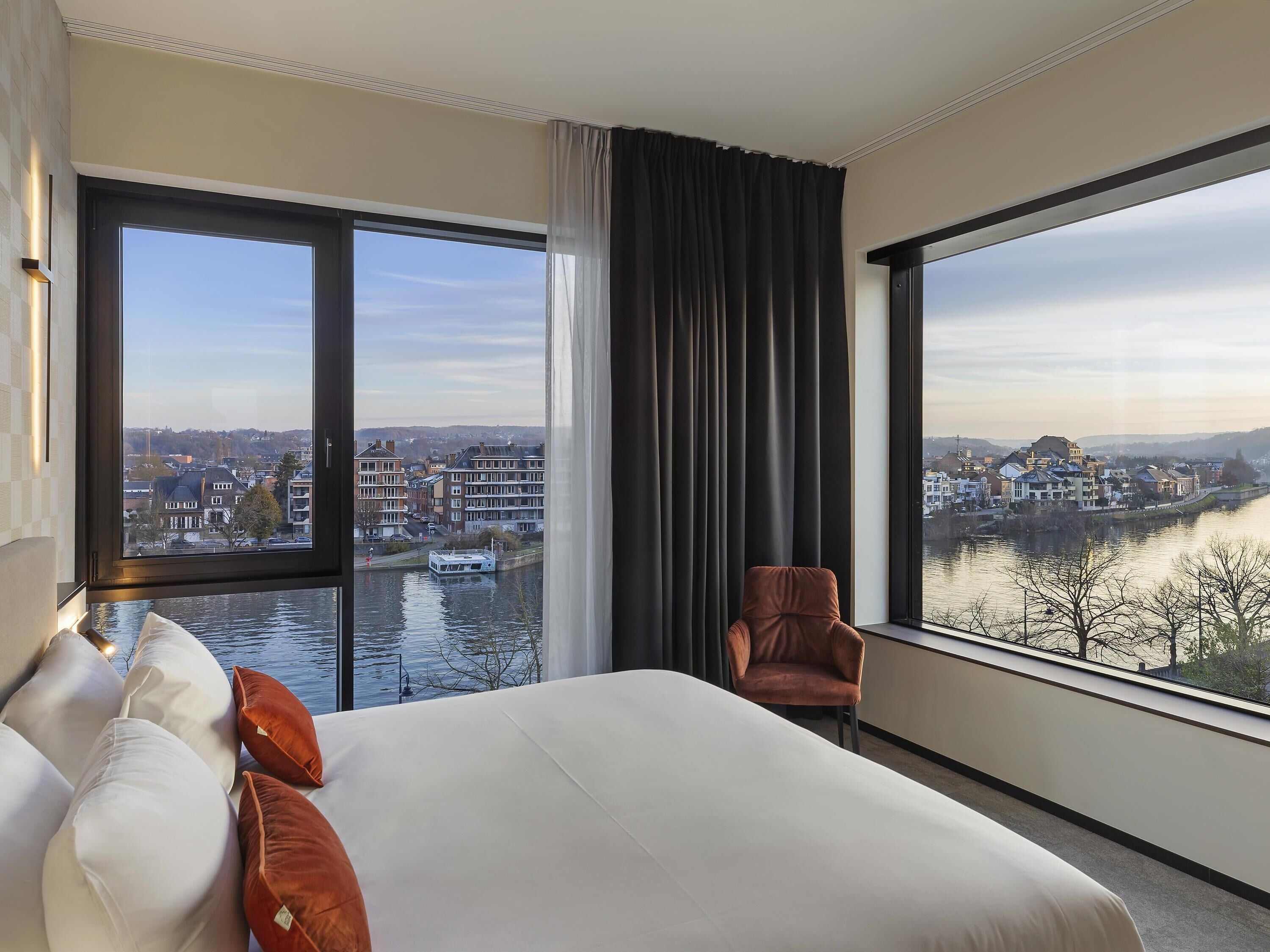 Une chambre d’hôtel avec un grand lit, une chaise et une vue sur une rivière et le paysage urbain, grâce à de grandes fenêtres.