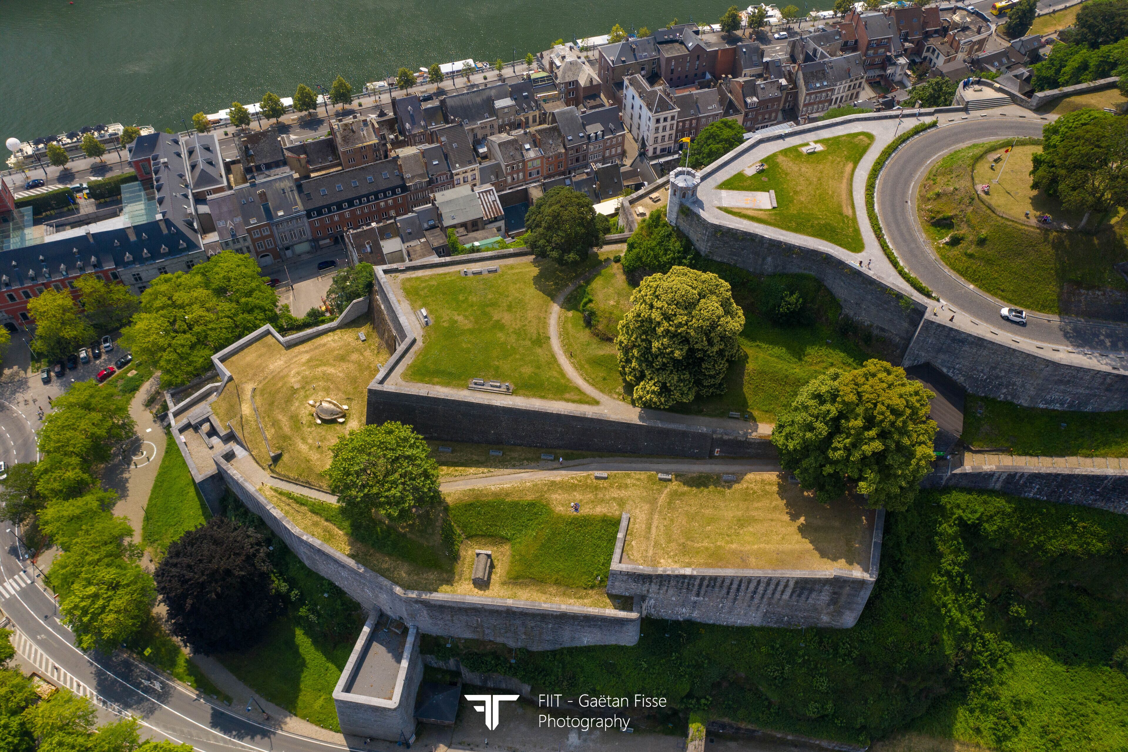 Vue aérienne d’une fortification historique, entourée de bâtiments urbains, avec une route sinueuse et une étendue d’eau.