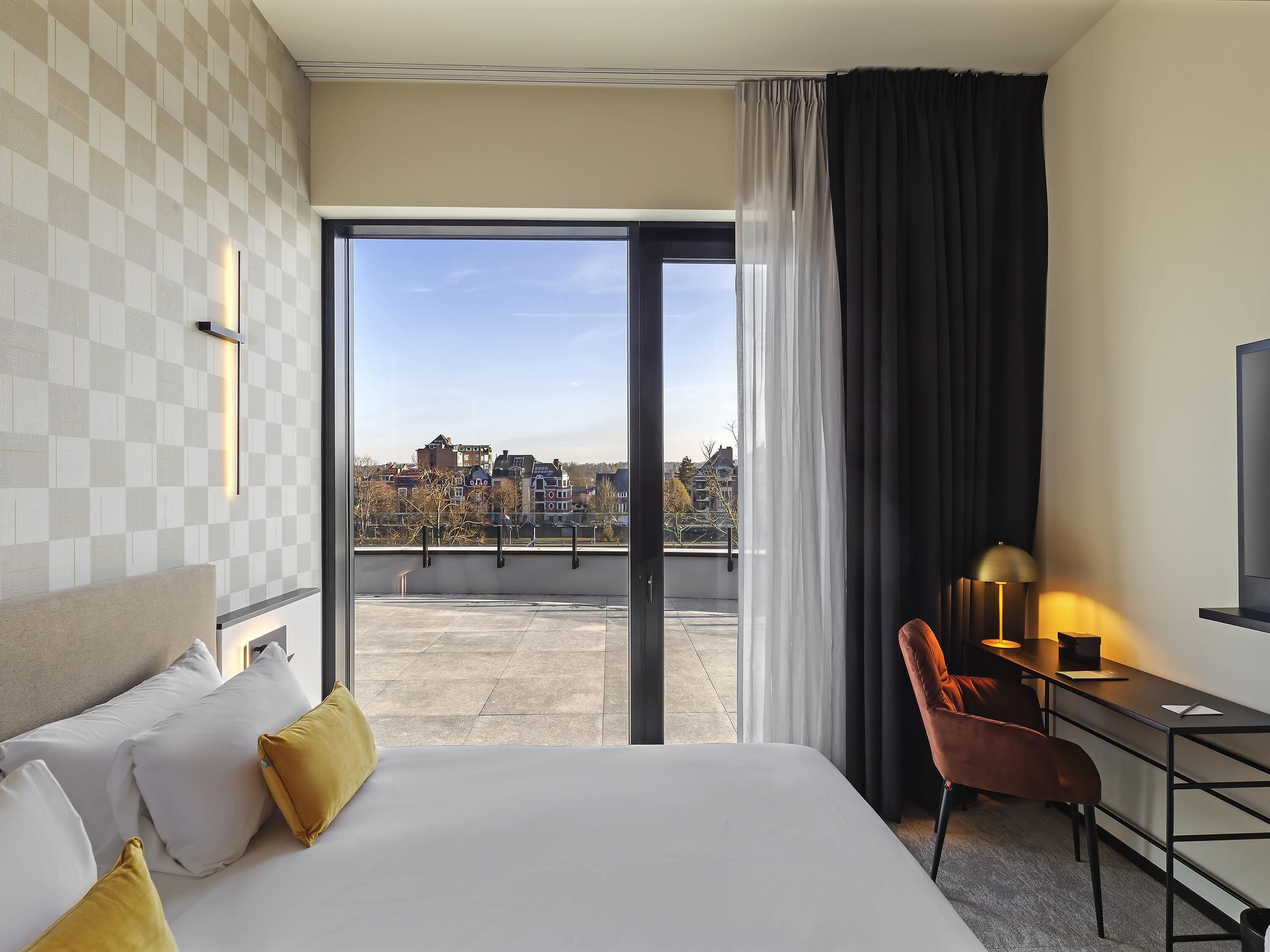 A hotel room with a large bed, a desk, a chair, and a view of the city through a large window.