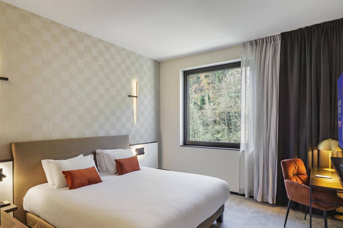 A hotel room with a large bed, a desk with a chair, a window with a view of trees, and a wall with a geometric pattern.