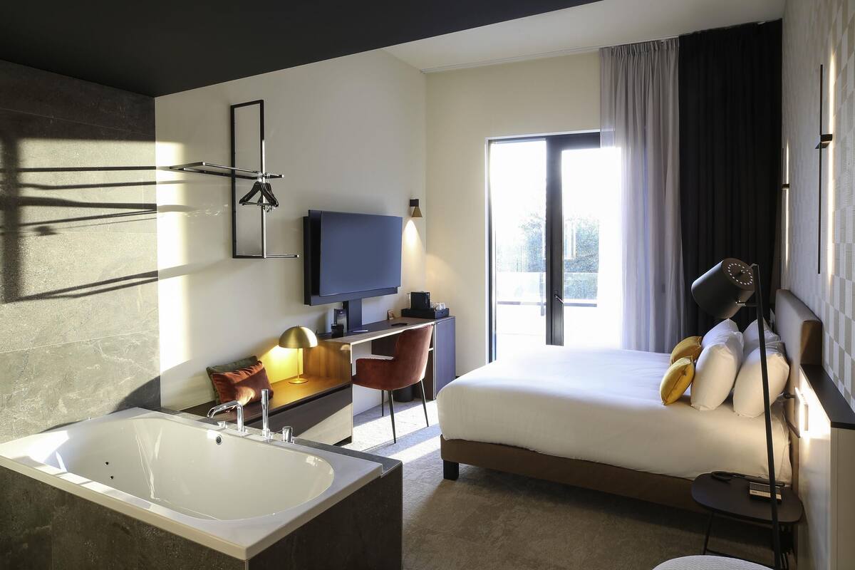 A modern hotel room with a large bed, a desk, a flat-screen TV, a bathtub, and a large window.
