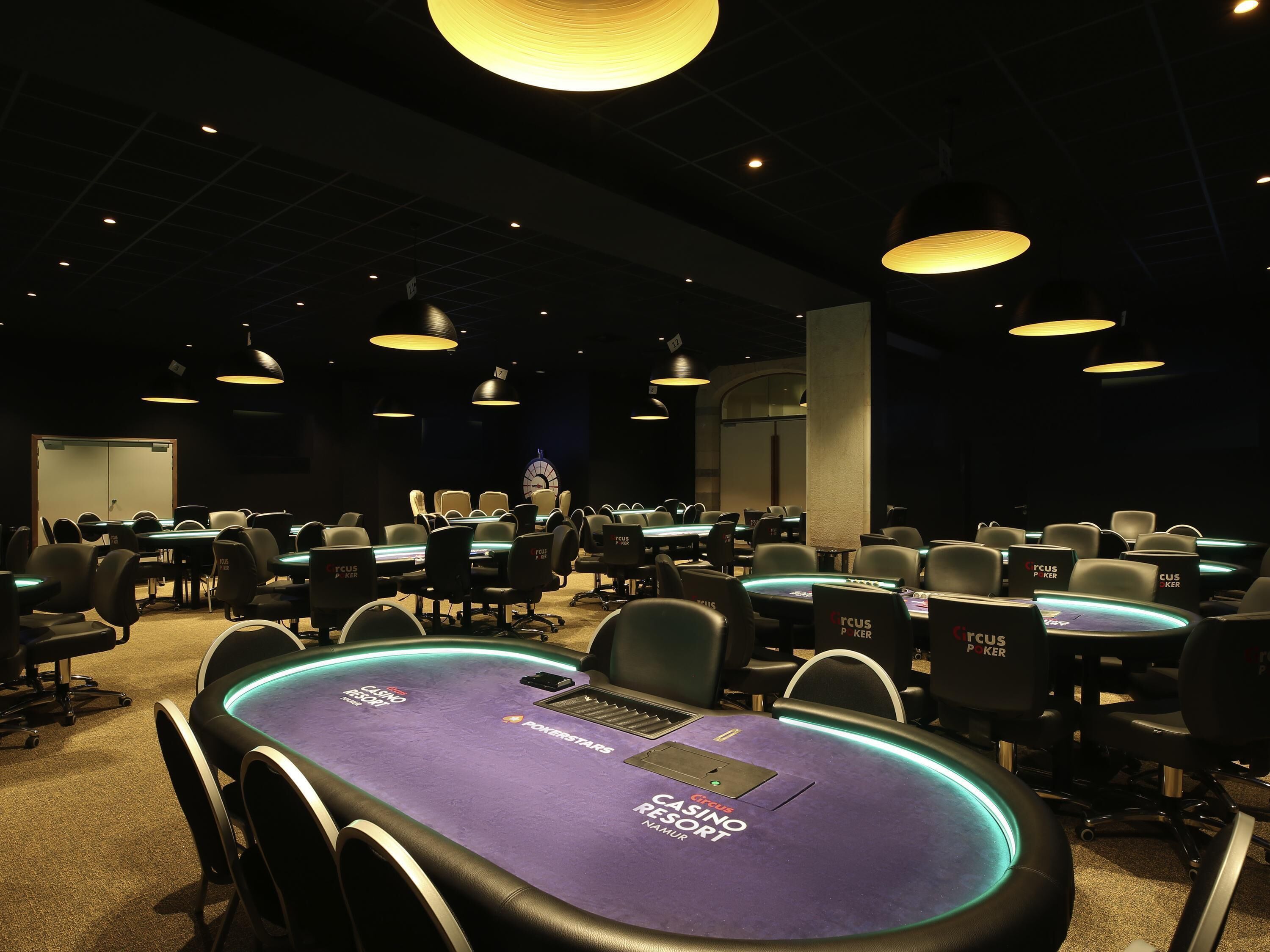 Een casino pokerruimte met meerdere tafels en stoelen, verlichting boven en een grote klok aan de muur.