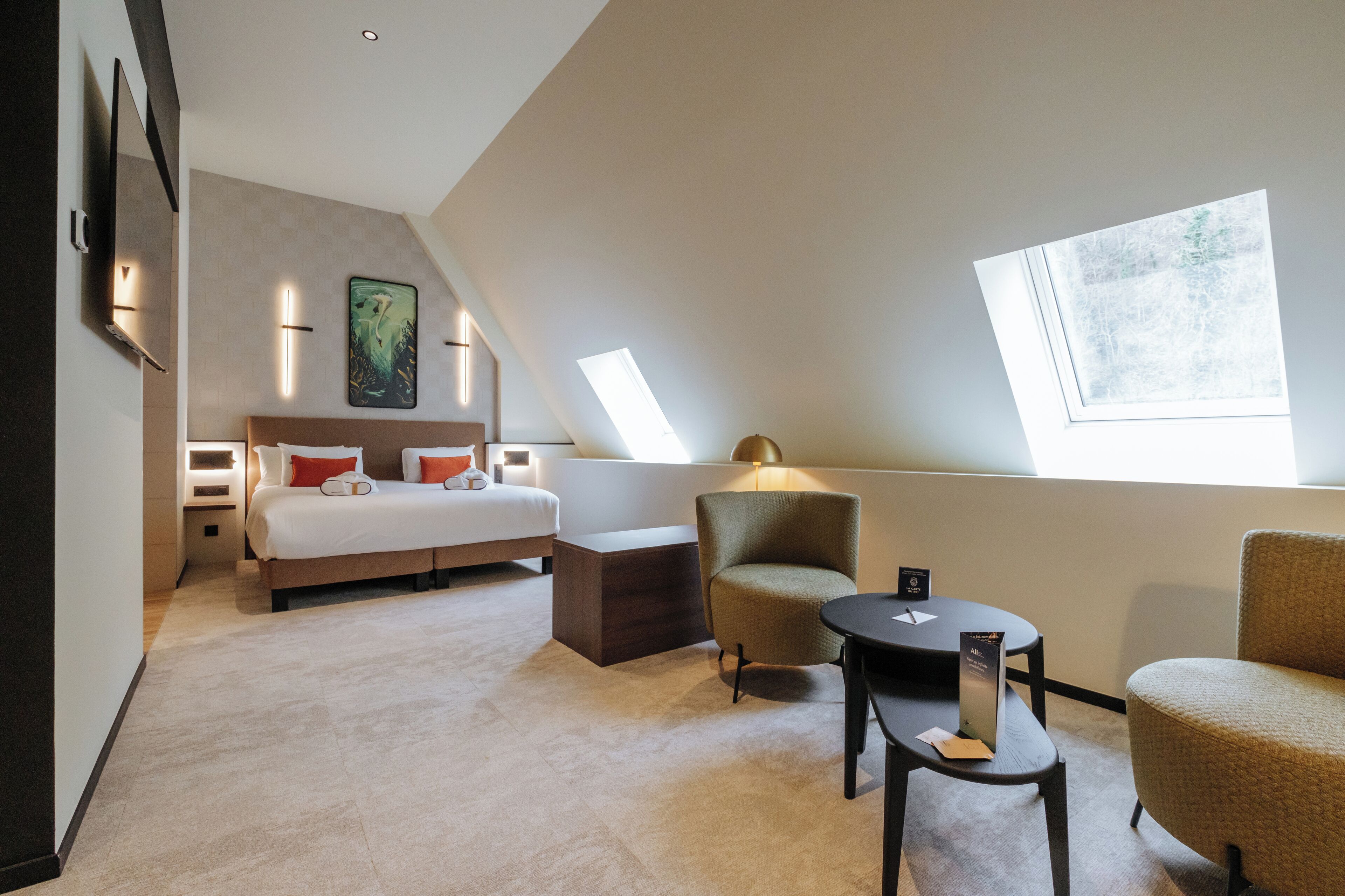 Una habitación de hotel moderna con una cama grande, una zona de estar con una silla y una mesa, un escritorio pequeño y un ventanal que proporciona luz natural.