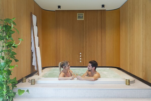 A couple in a hot tub with wooden walls and a plant.