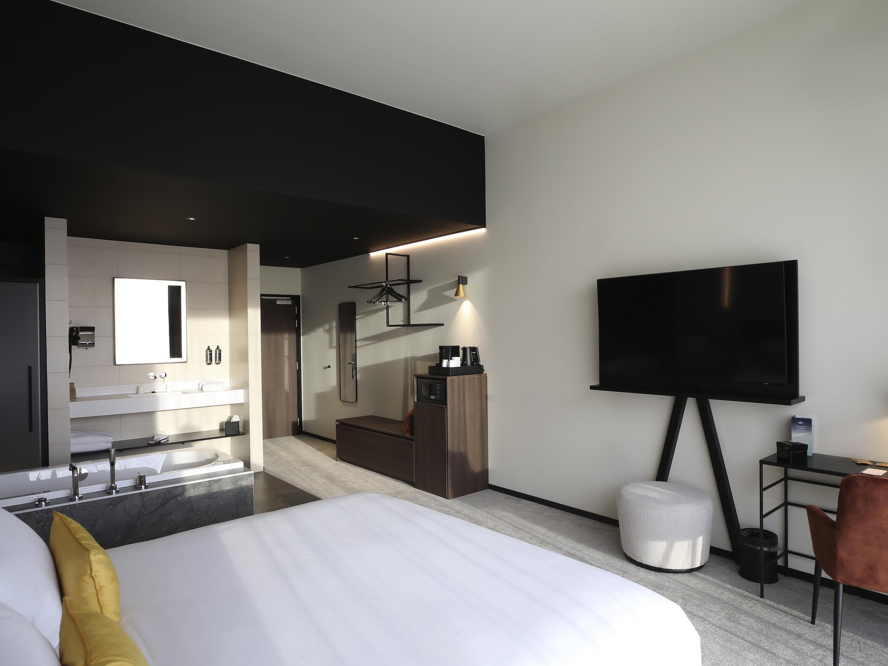 A modern hotel room with a large bed, a TV, a desk with a chair, a bathroom with a mirror, and a kitchenette.