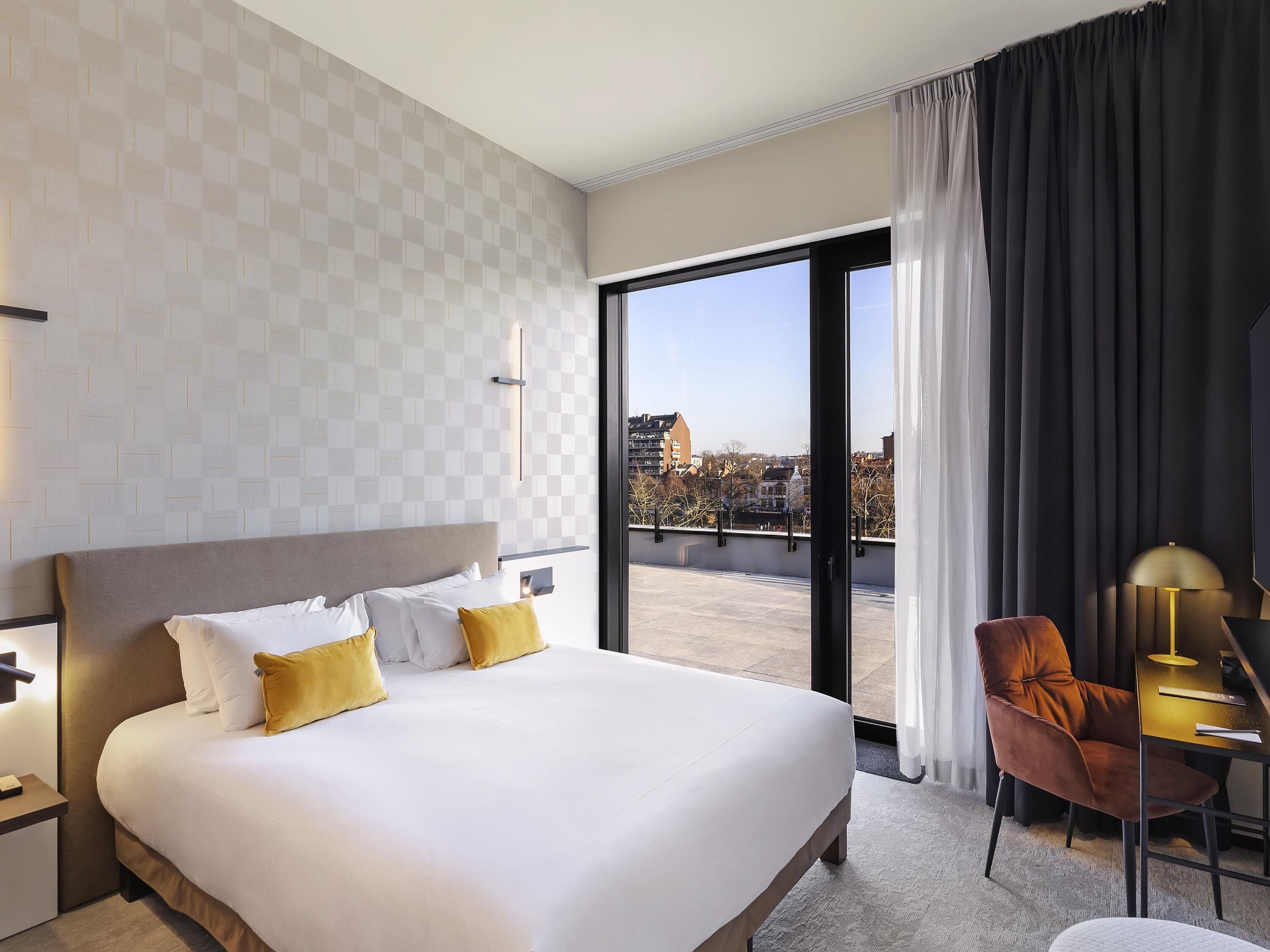 Ein Hotelzimmer mit einem großen Bett, einem Schreibtisch mit Stuhl, Blick auf die Stadt durch ein großes Fenster und einem modernen Design.