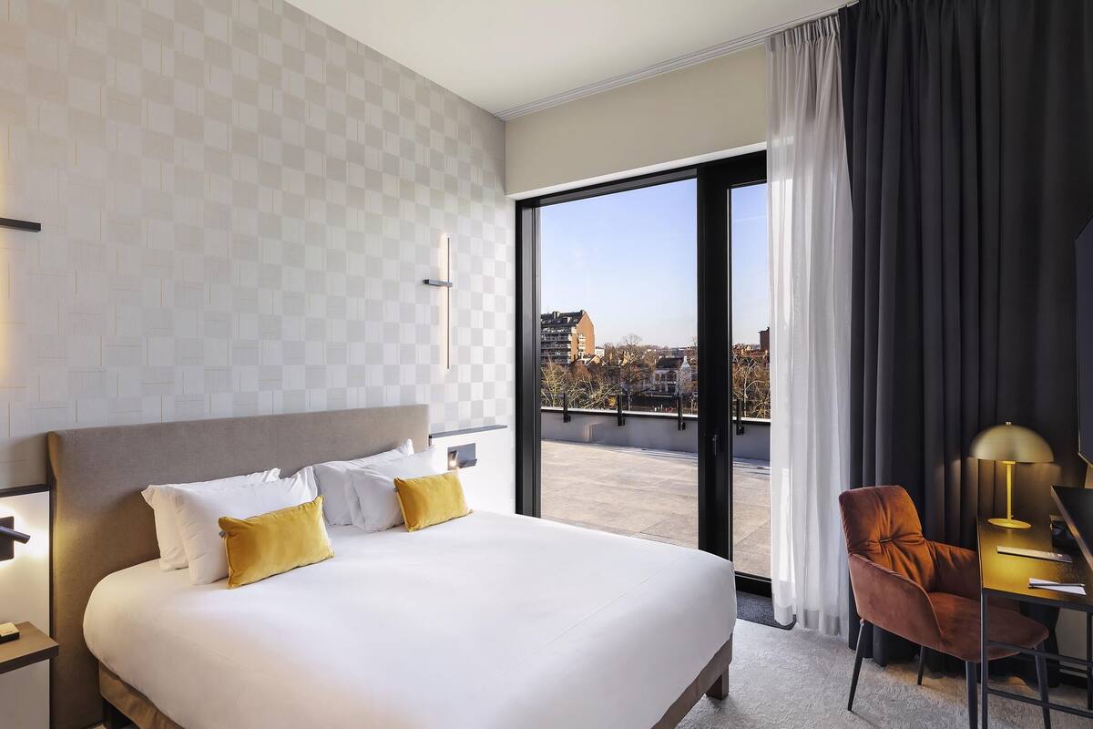 A hotel room with a large bed, a desk with a chair, a view of the city through a large window, and a modern design.