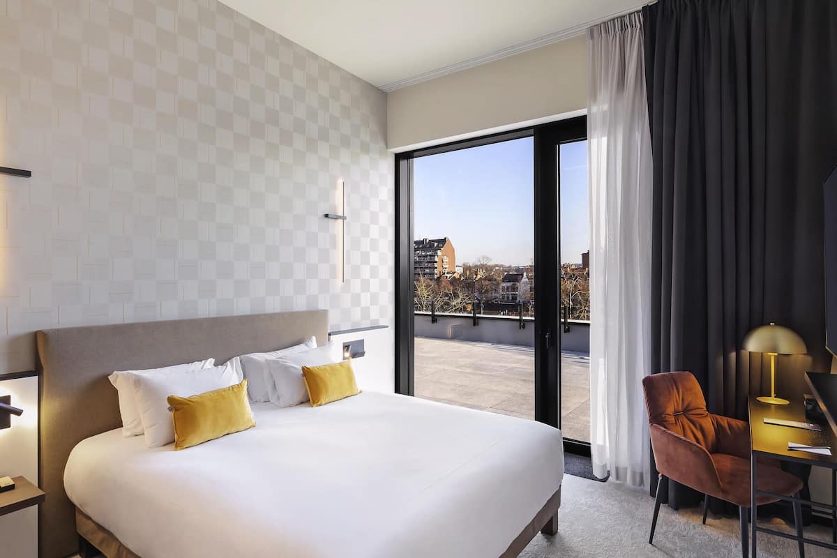 Ein Hotelzimmer mit einem großen Bett, einem Schreibtisch mit Stuhl, Blick auf die Stadt durch ein großes Fenster und einem modernen Design.
