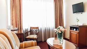 Une chambre d’hôtel avec un canapé, un fauteuil, une table ornée d’un vase de fleurs, un téléviseur fixé au mur et un bureau avec une lampe et une tasse.