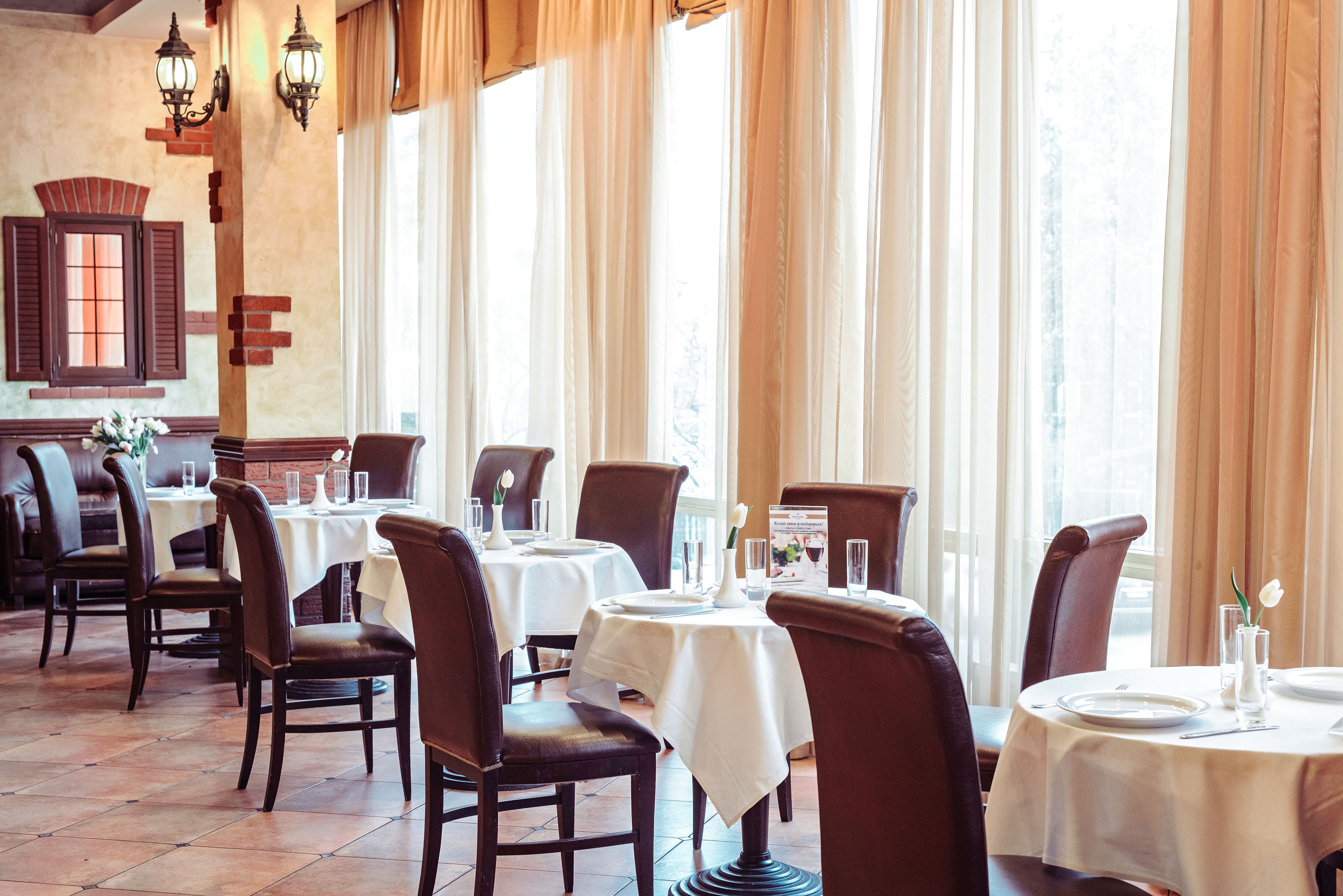 Un restaurante con varias mesas y sillas, ventanas amplias con cortinas y paredes de ladrillo.