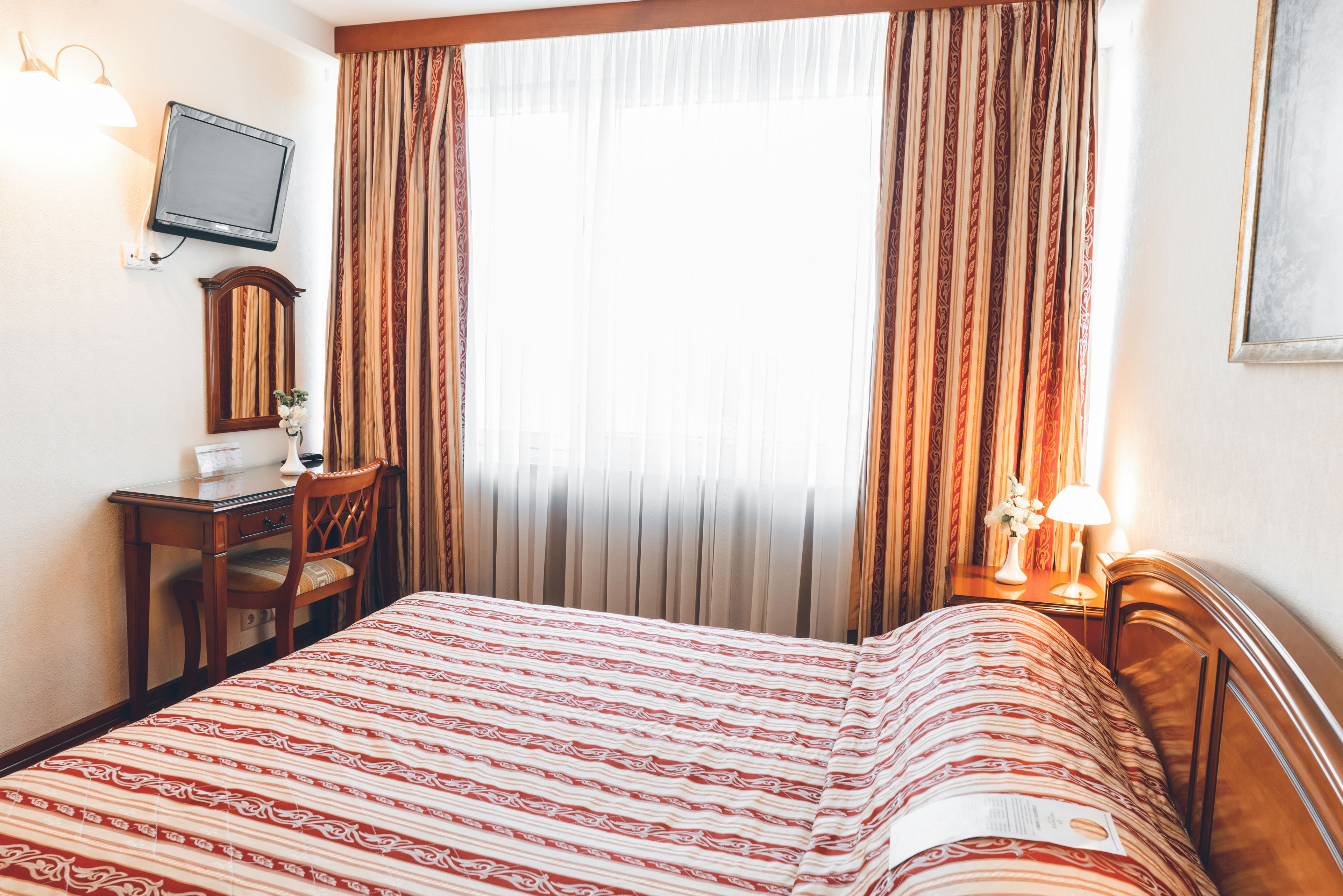 Quarto de hotel com cama, escrivaninha, cadeira, televisão e cortinas.