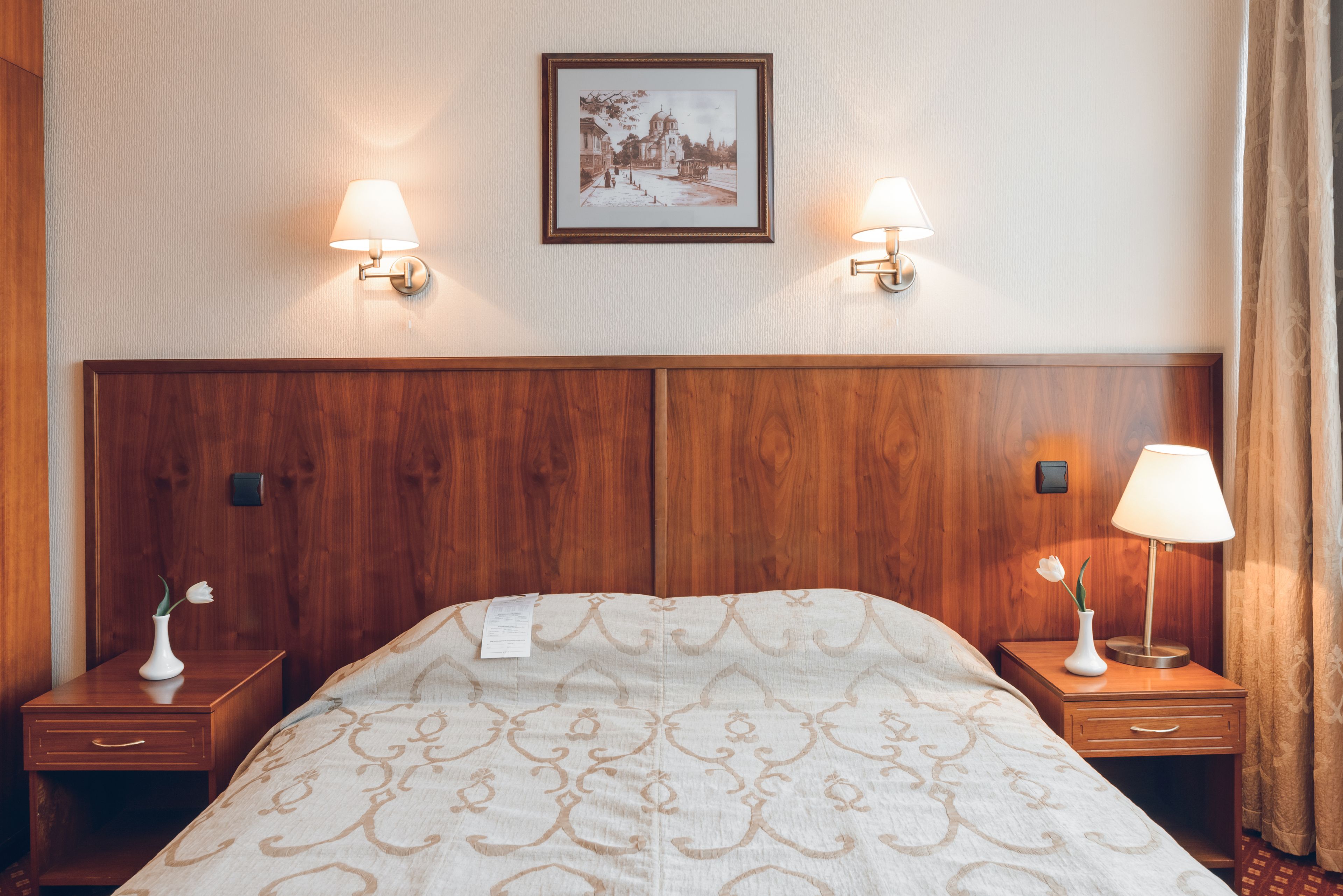 Habitación de hotel con una cama grande, cabecera de madera, dos mesitas de noche con lámparas, un cuadro enmarcado en la pared y un pequeño florero con una flor sobre una de las mesitas.