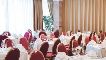 Een balzaal met gedekte tafels, rode fauteuils en een groot raam met gordijnen.
