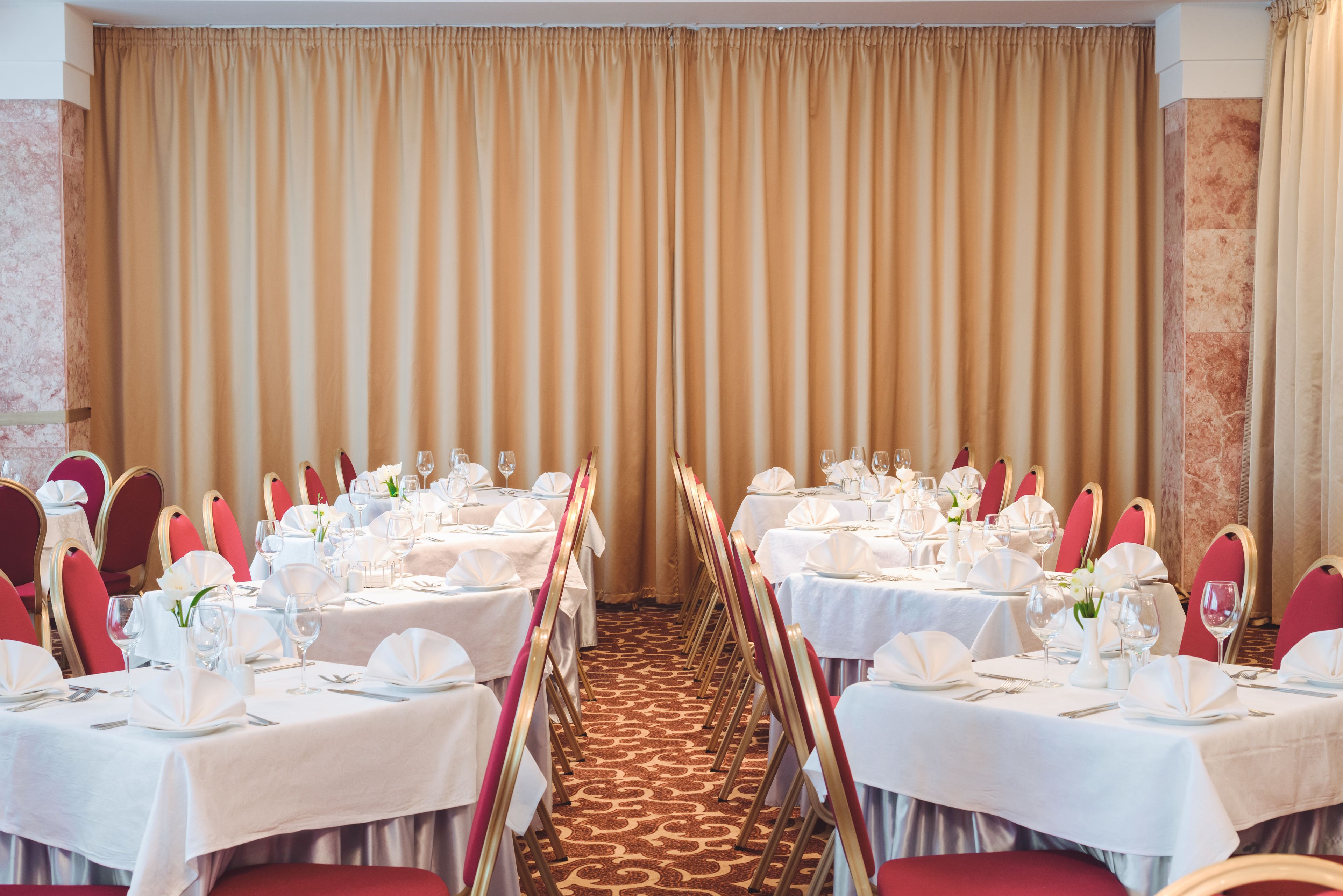 Uma sala de banquetes com mesas montadas para um evento formal, com toalhas brancas, cadeiras vermelhas e piso coberto por carpete.