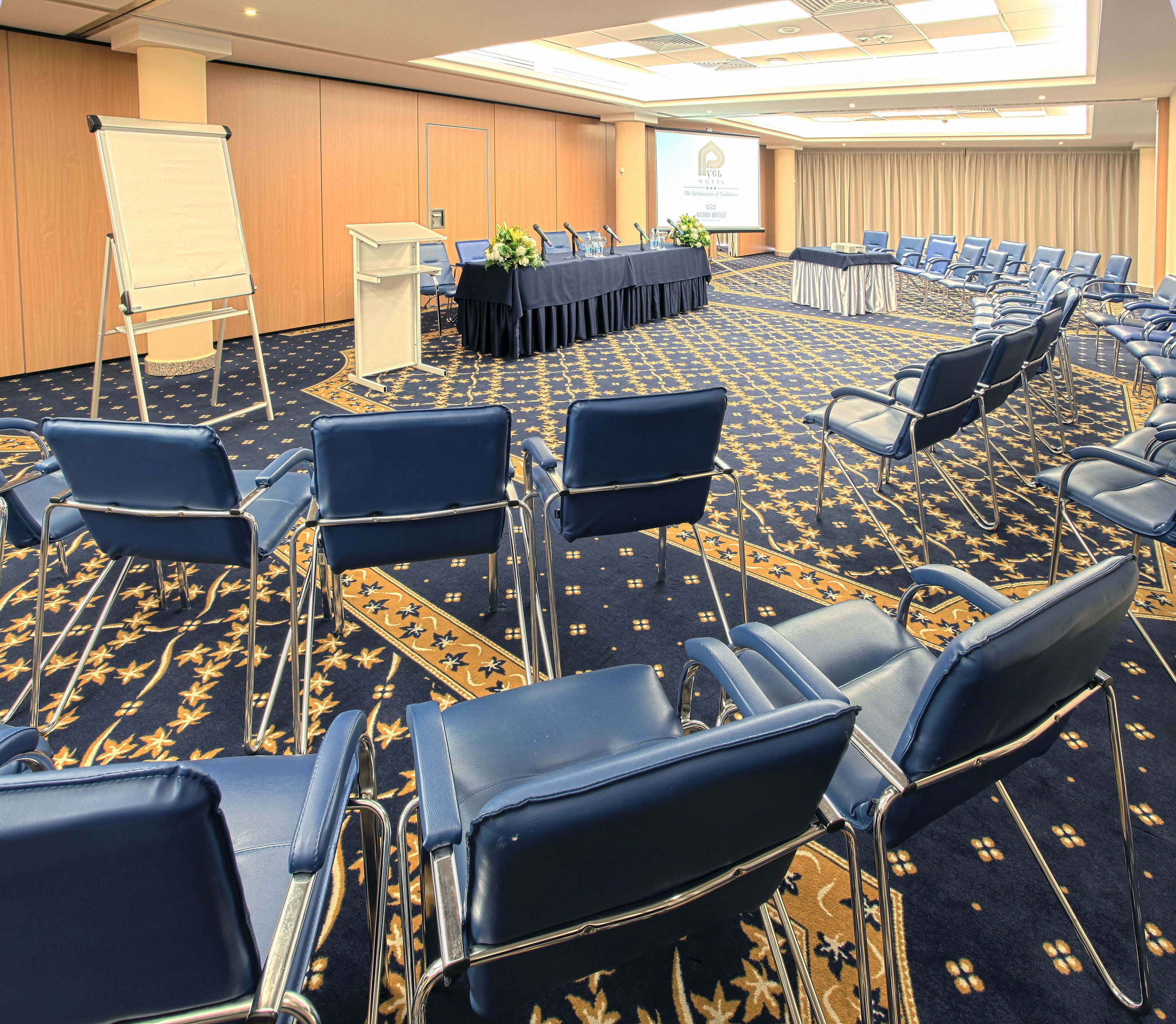 A conference room with rows of blue chairs, a whiteboard, a podium, and a projector screen.