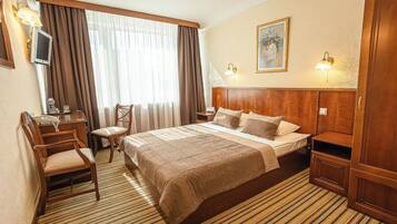Habitación de hotel con cama, escritorio, silla, televisor y ventana con cortinas.