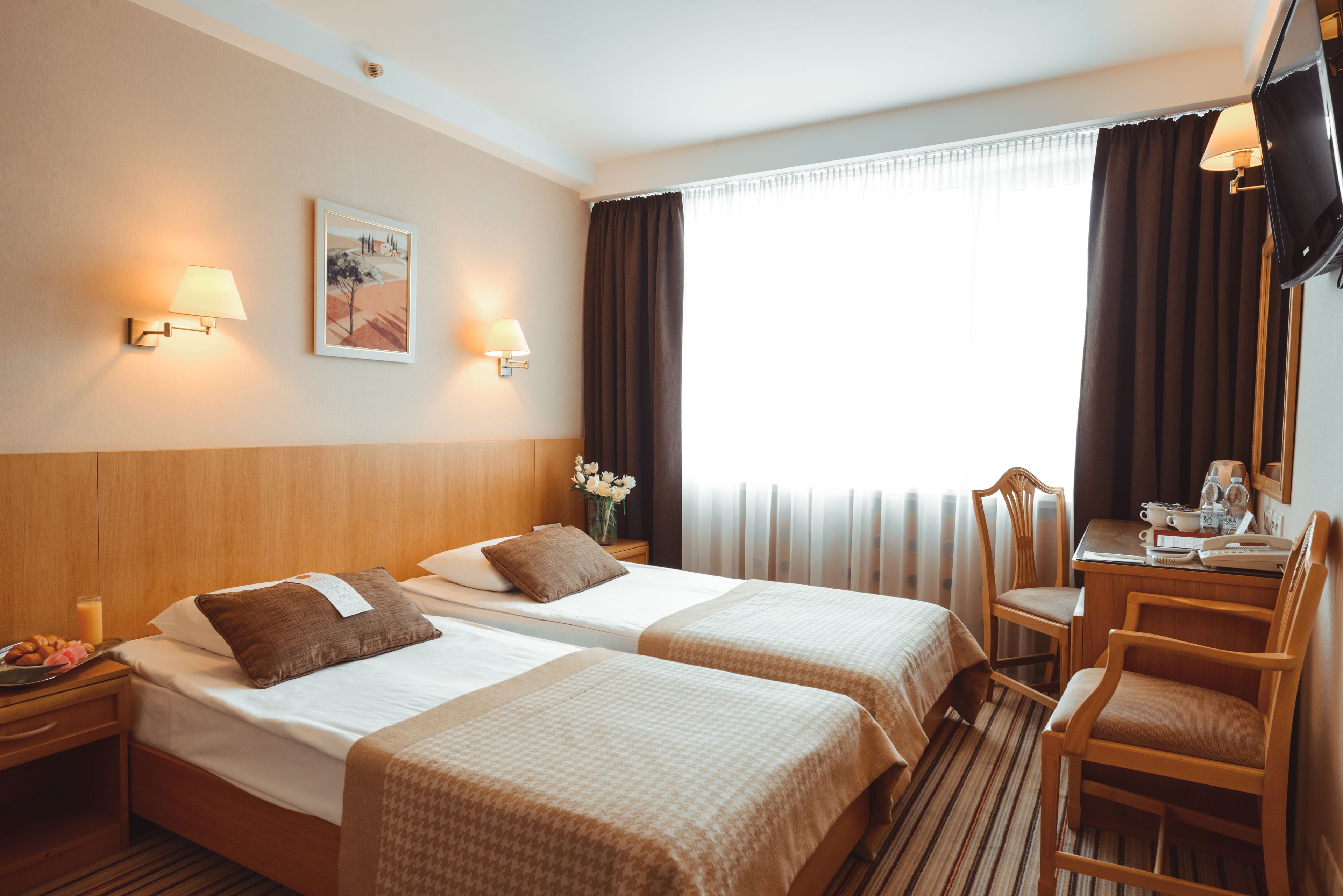 A hotel room with two beds, a desk with a chair, a TV, and a window with curtains.