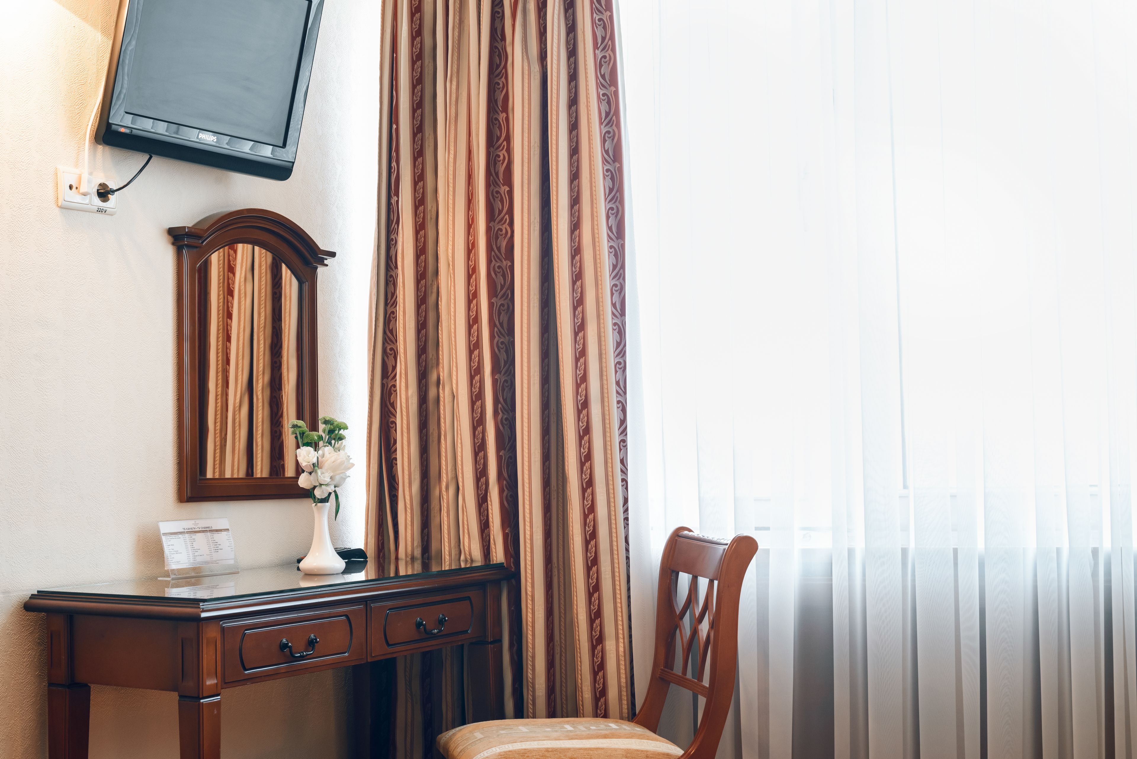 Una camera d'albergo con una scrivania in legno, uno specchio, una TV a schermo piatto montata a parete, una sedia e una finestra con tende leggere.