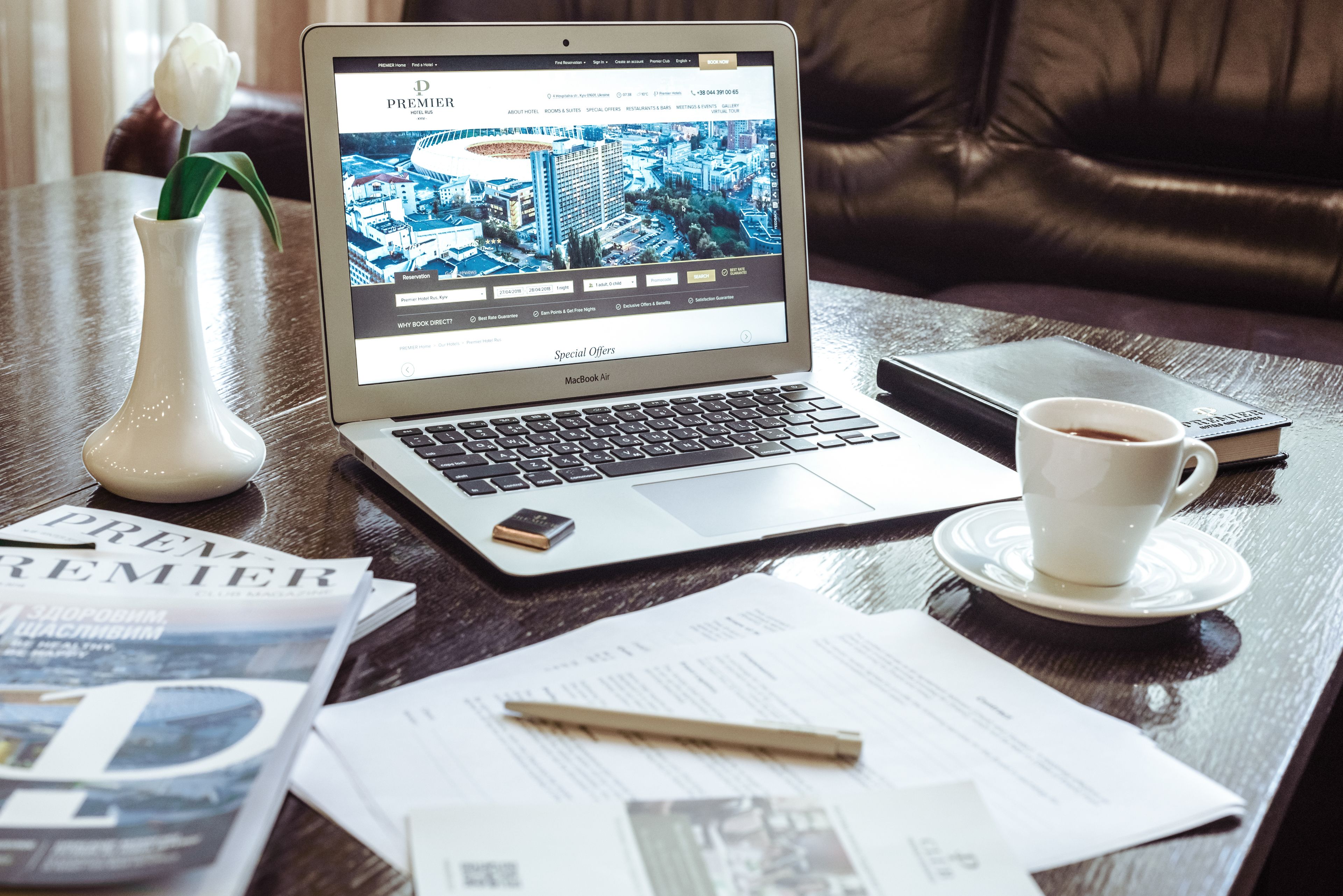 A laptop on a wooden table displaying a hotel website, a white vase with a flower, a cup of coffee, and some documents.