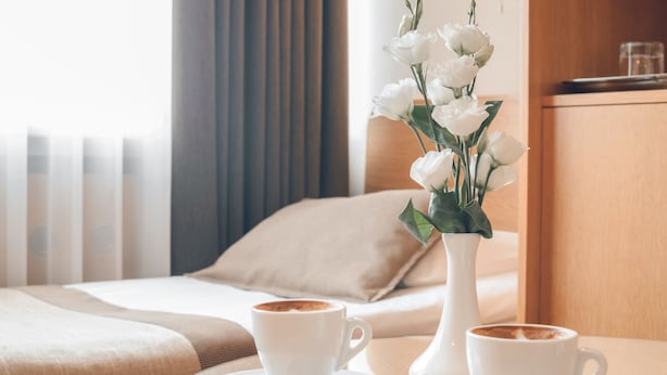 Une chambre d’hôtel avec un lit, deux tasses de café sur un plateau et un vase rempli de fleurs blanches.