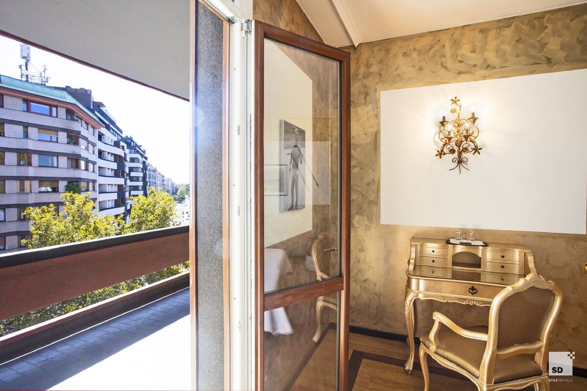 A room with a view of a building and trees, a glass door, a wall-mounted light fixture, and a wooden desk with drawers.