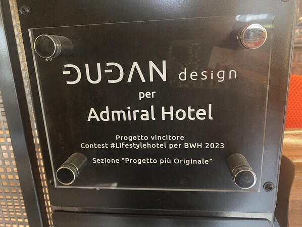 A plaque on a door with text indicating a design contest for the Admiral Hotel, part of a lifestyle hotel contest for BWH 2023, and a section for the most original project. - Admiral Hotel (Milan)