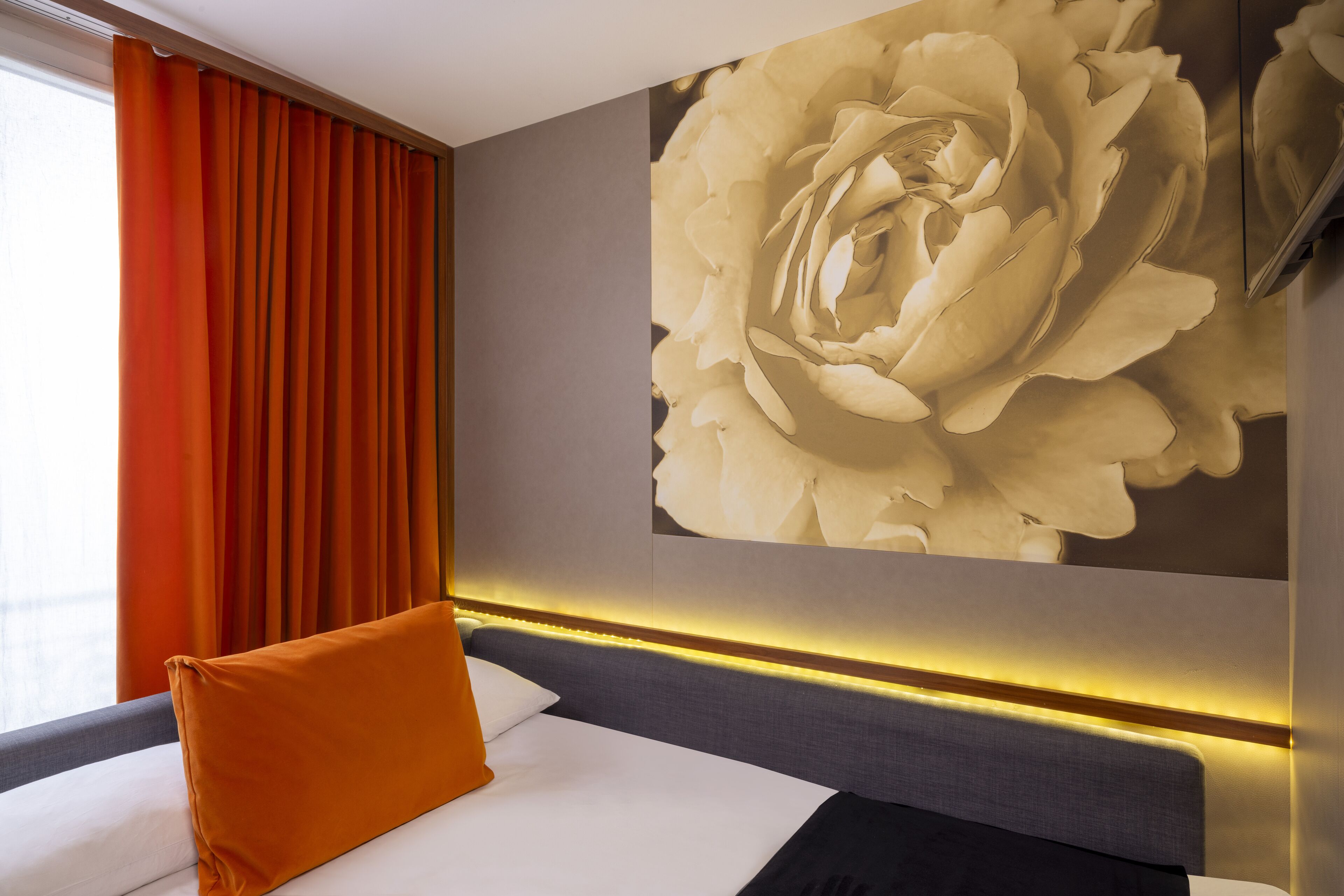 A hotel room with a large floral artwork on the wall, a bed with white bedding, an orange pillow, and a grey headboard.