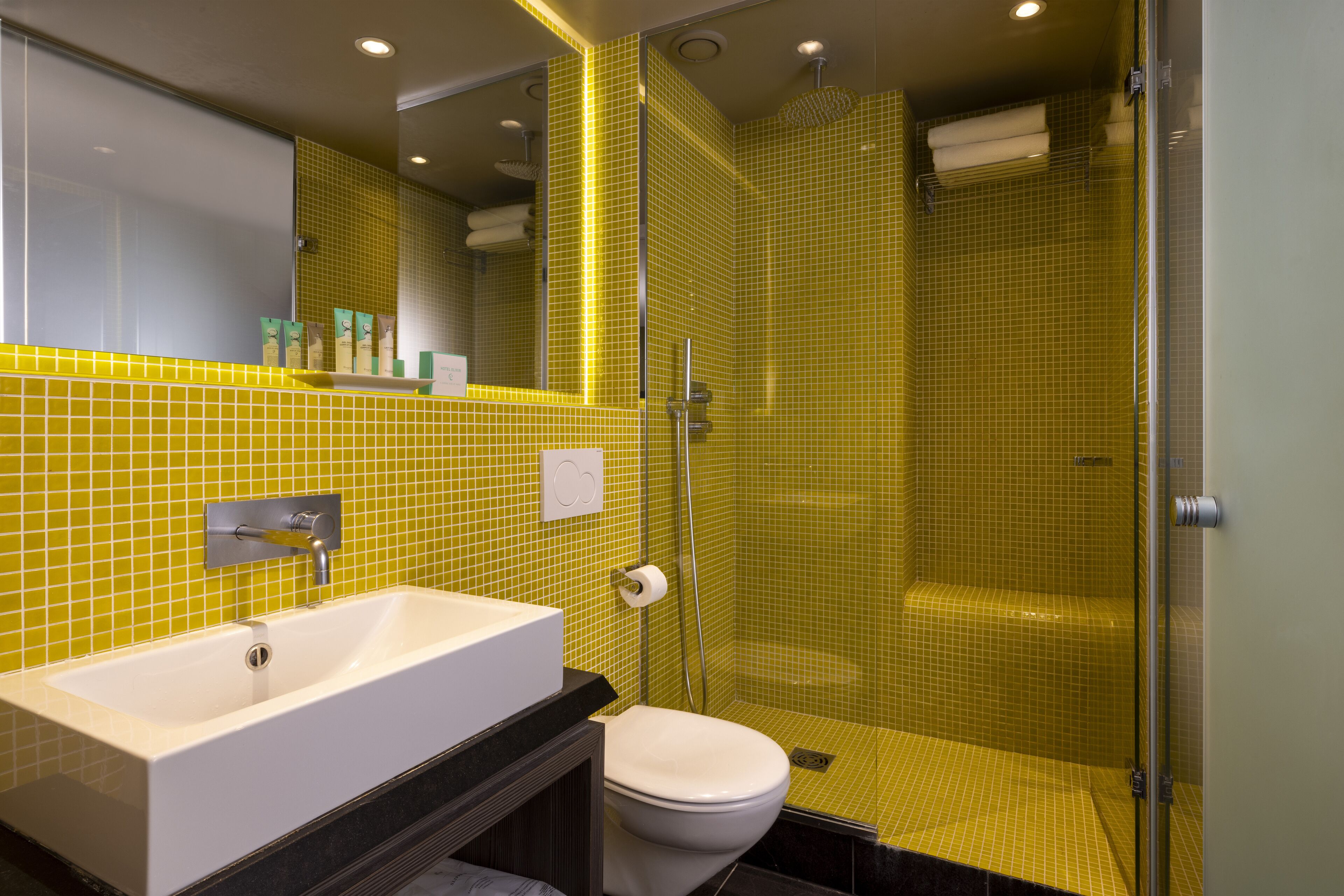 A bathroom with a white sink, toilet, and shower area, featuring yellow-tiled walls and a glass shower enclosure.