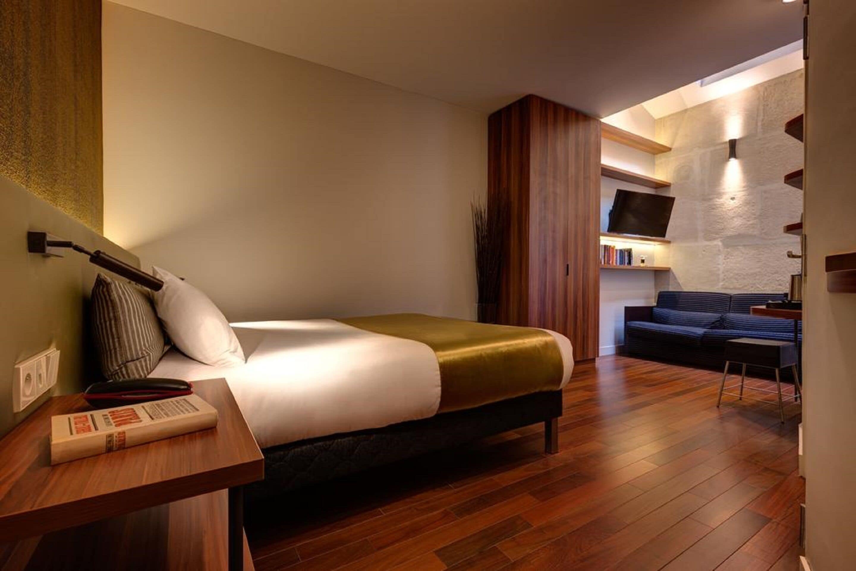 Una camera da letto moderna con un letto grande, un comodino in legno con un libro, un divano scuro e una TV montata su una parete in pietra.