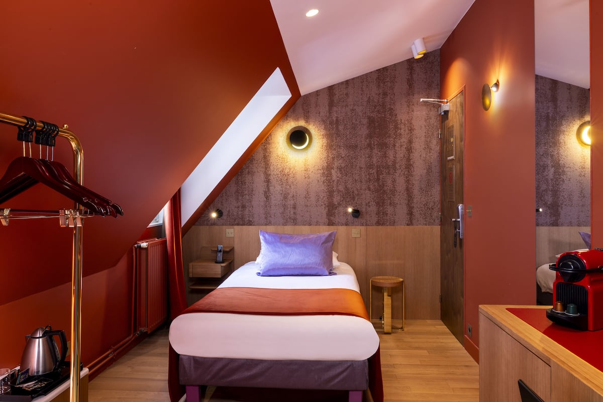 Ein modernes Hotelzimmer mit schrägverdeckter Decke, einem Bett mit violettem Kopfkissen, einer Kaffeemaschine und einem Holznachttisch.