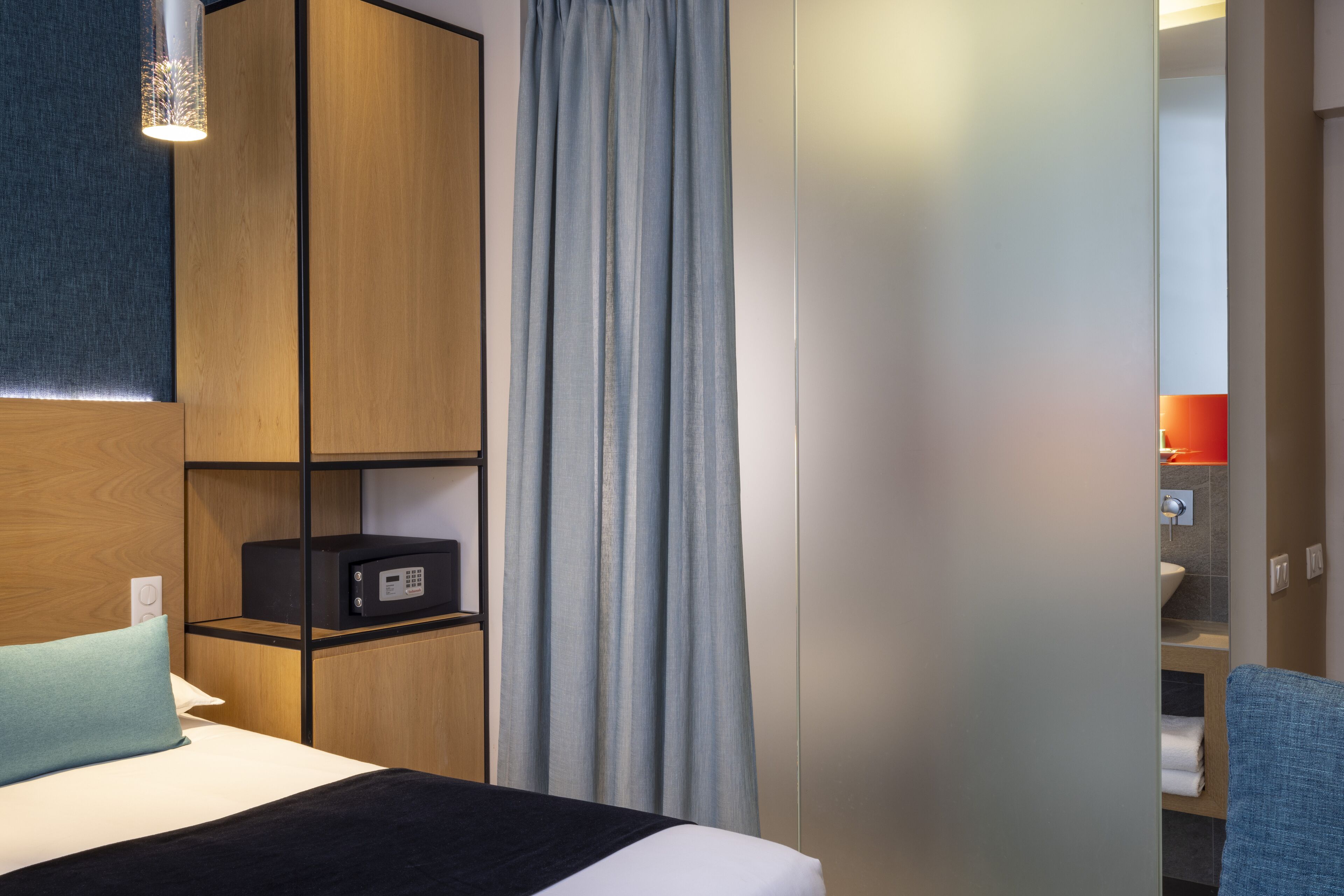 A hotel room with a bed, a wooden wardrobe, a desk with a computer, and a bathroom visible through an open door.
