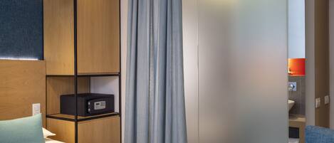 A hotel room with a bed, a wooden wardrobe, a desk with a computer, and a bathroom visible through an open door.