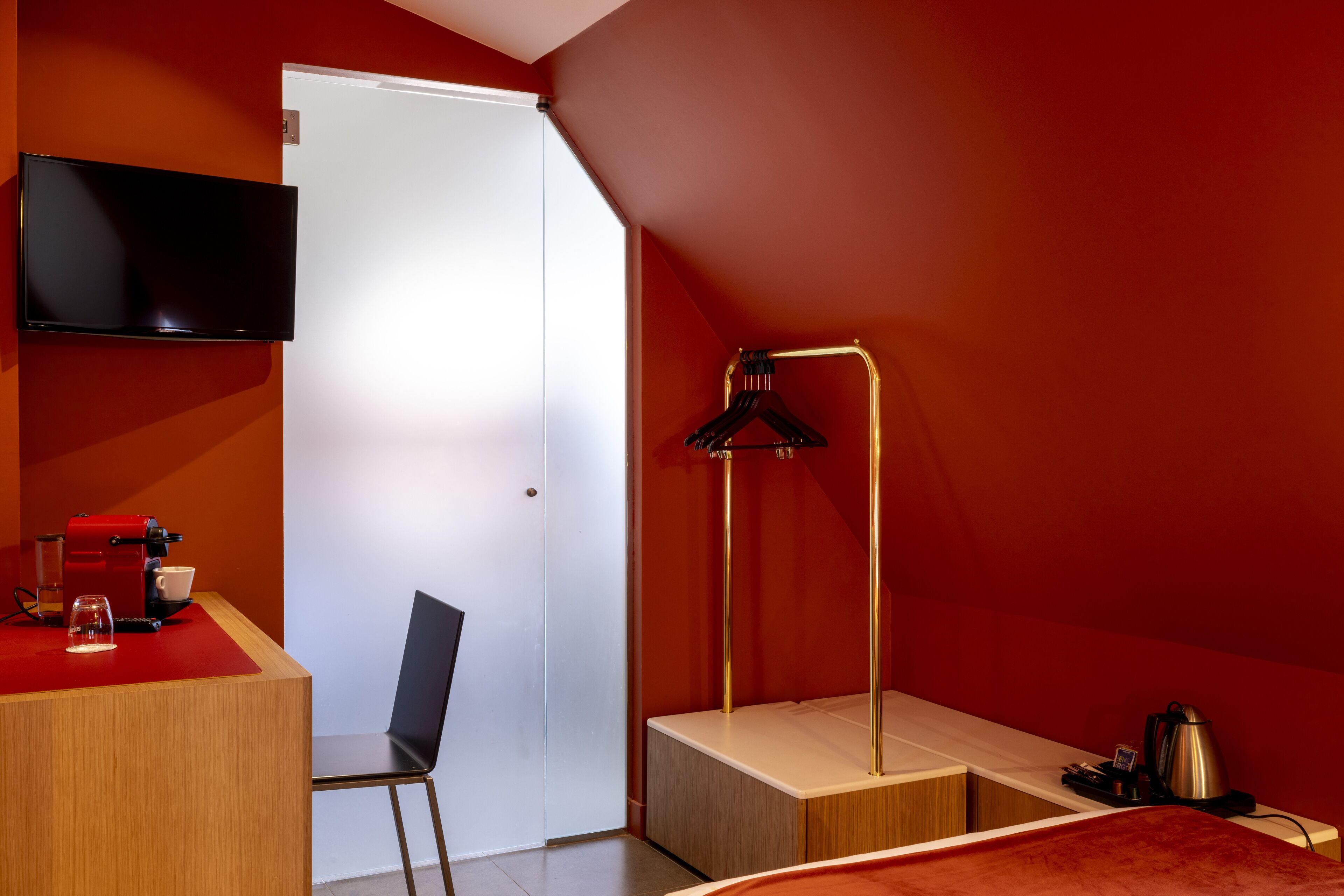 A compact hotel room with a flat-screen TV mounted on the wall, a desk with a coffee machine, a chair, and a bed with a red blanket.
