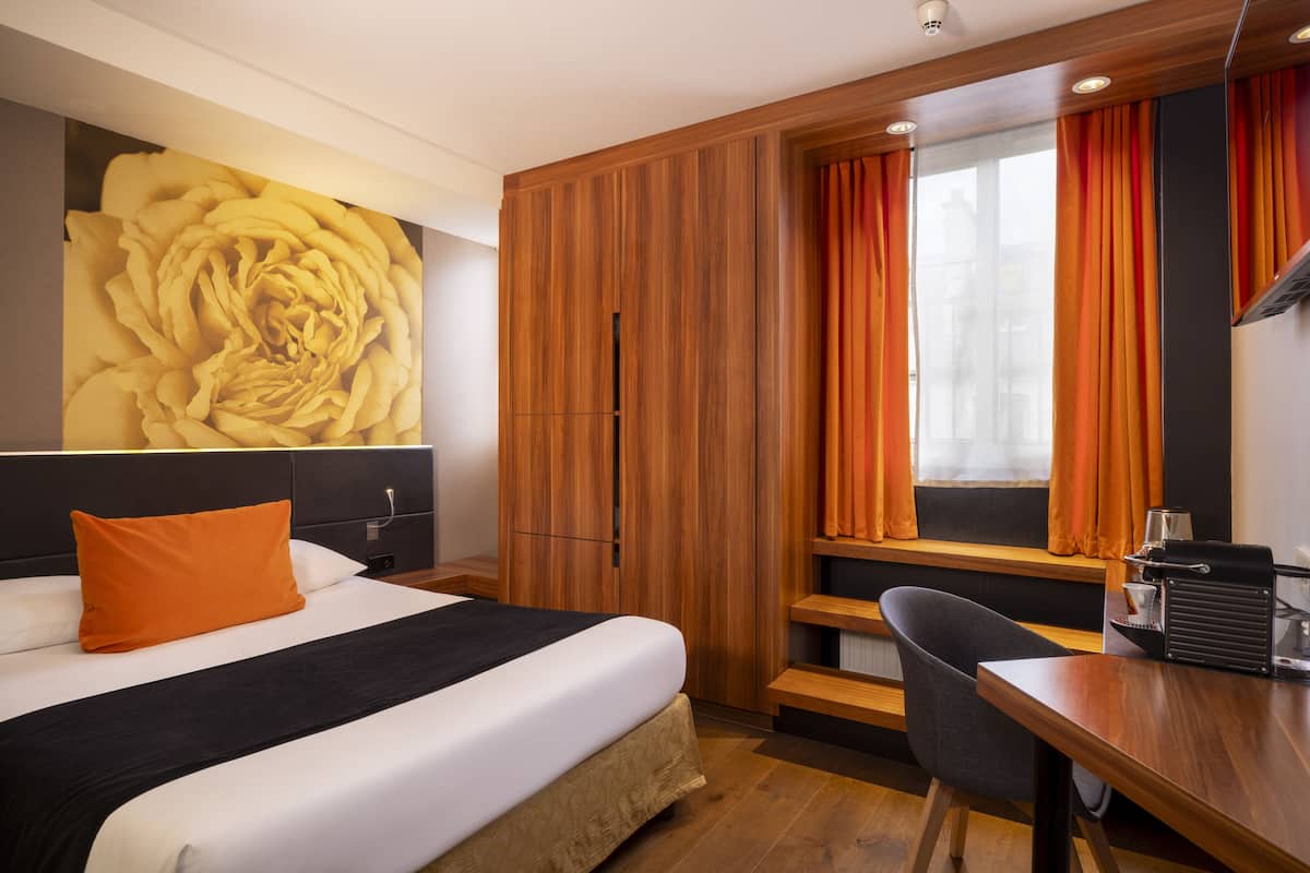 Ein Hotelzimmer mit einem großen Bett, einem Schreibtisch, einem Stuhl, einem Fenster mit Vorhängen und einem Wandbild mit floralem Muster.