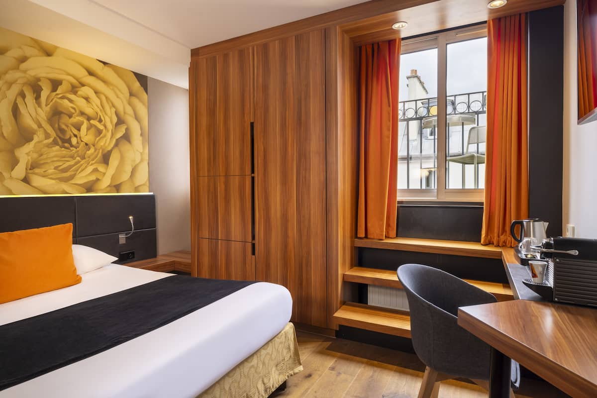 Ein Hotelzimmer mit einem Bett, einem Holzschrank, einem Schreibtisch mit Stuhl, einem Fenster mit Vorhängen und einem großen Blumenbild an der Wand.