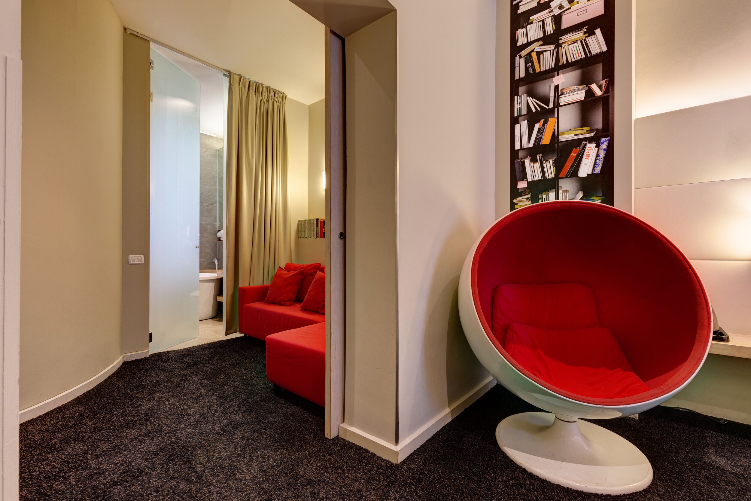 A modern hotel room with a red chair, a bookshelf, and a red sofa.