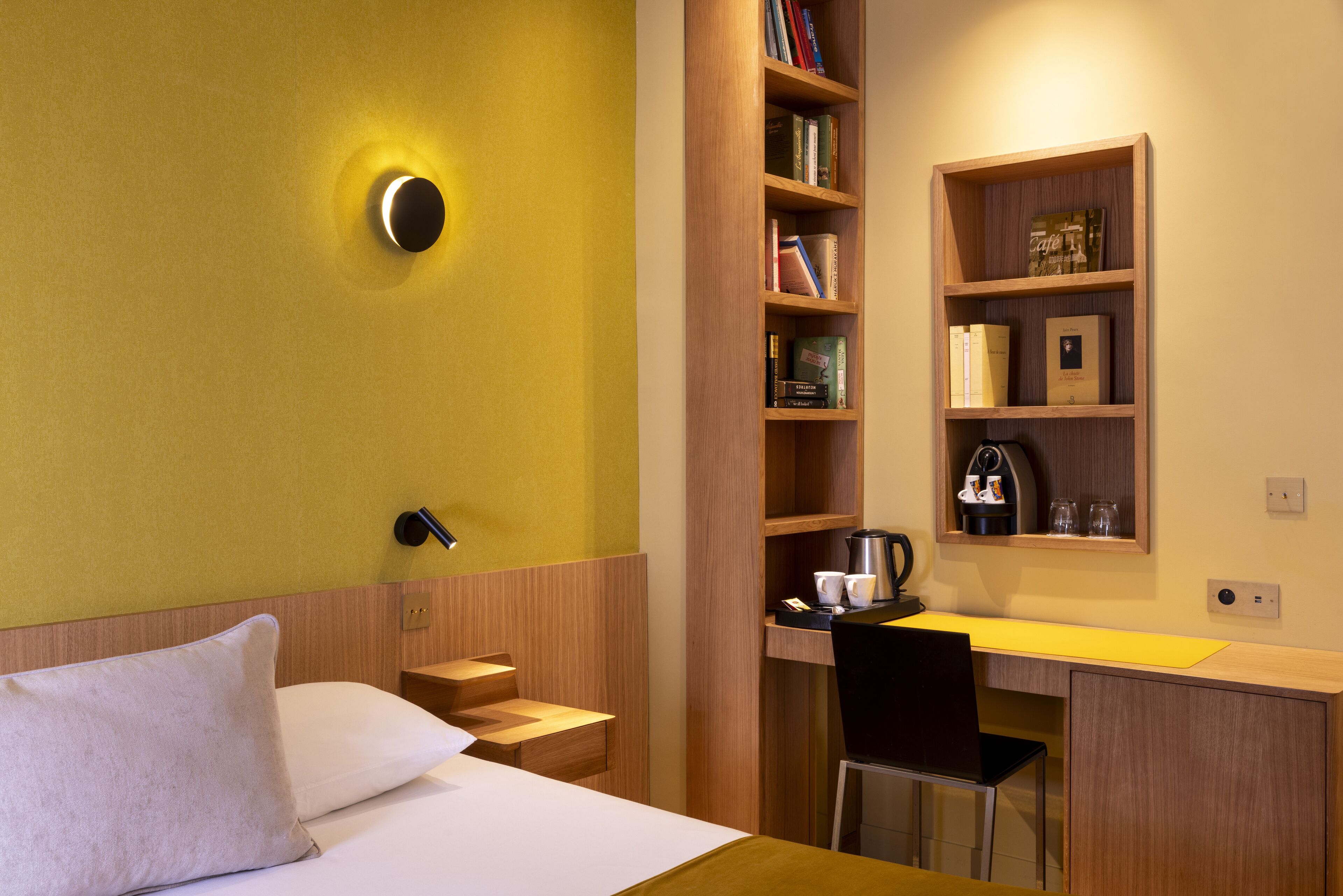 A hotel room with a bed, a desk, a chair, a bookshelf, and a coffee machine.