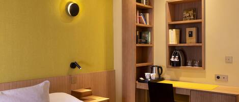 A hotel room with a bed, a desk, a chair, a bookshelf, and a coffee machine.