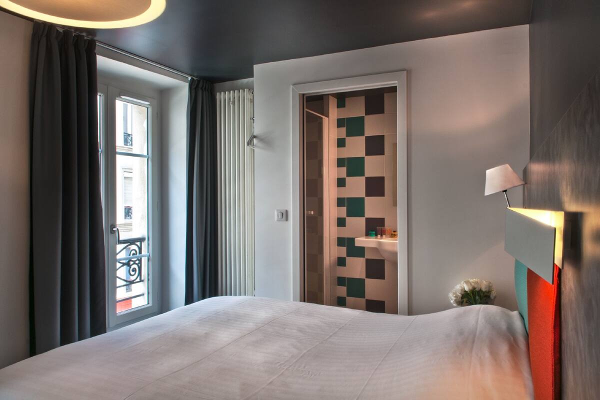 A hotel room with a bed, a door with a checkered pattern, a window with curtains, and a wall-mounted lamp.