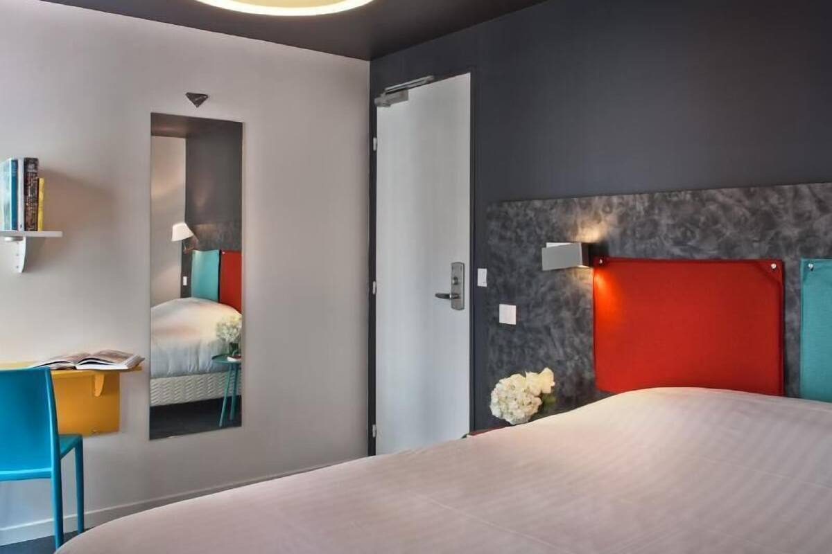 A modern hotel room with a bed, a desk, a chair, and a door.