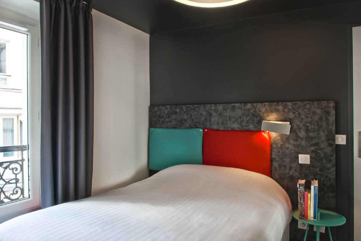 A modern hotel room with a large bed, a headboard with teal and red cushions, a small side table with books, and a window with curtains.