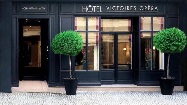 L’extérieur de l’hôtel Victoires Opéra se distingue par ses ornements noirs, ses grandes fenêtres et les deux arbres taillés en forme de cône qui se trouvent devant l’entrée.