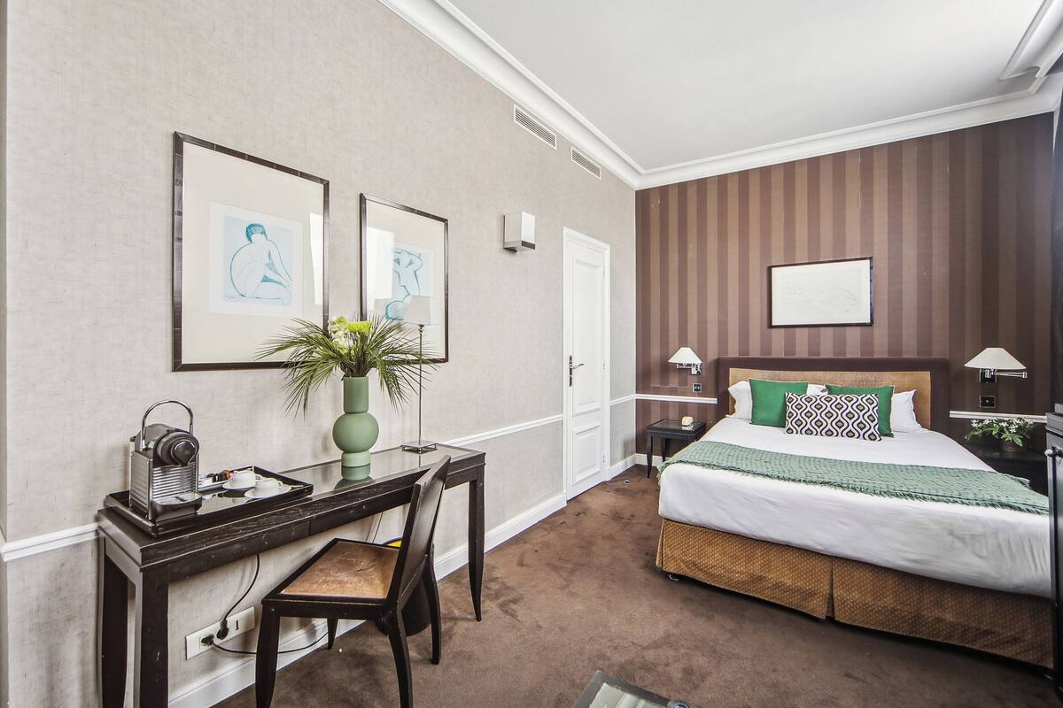 A hotel room with a bed, a desk with a chair, a small table with a plant, and framed artwork on the walls.