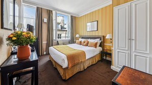 Une chambre d’hôtel avec un grand lit, une table de chevet, un bureau avec une lampe, une plante en pot et une vue sur la ville.