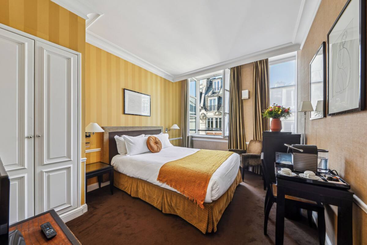 A hotel room with a large bed, a desk, a chair, a window with a view of buildings, and a potted plant.