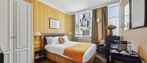 A hotel room with a large bed, a desk, a chair, a window with a view of buildings, and a potted plant.