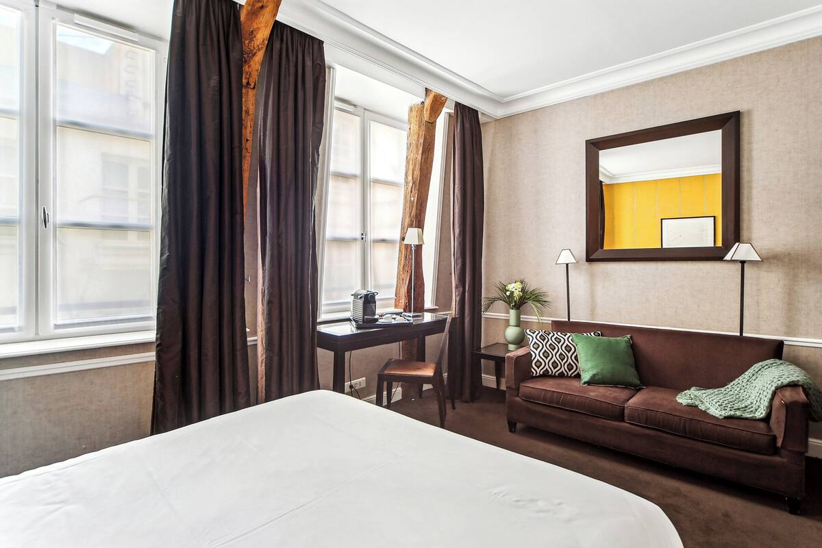 A hotel room with a bed, a brown leather sofa, a desk with a chair, and a large mirror on the wall.