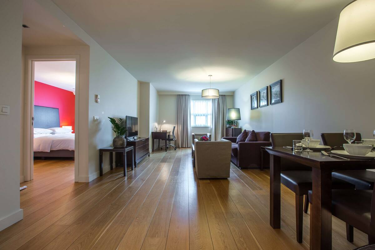 Una habitación de hotel moderna con piso de madera, una zona de comedor con una mesa dispuesta para dos personas, un sofá y un televisor en la esquina.