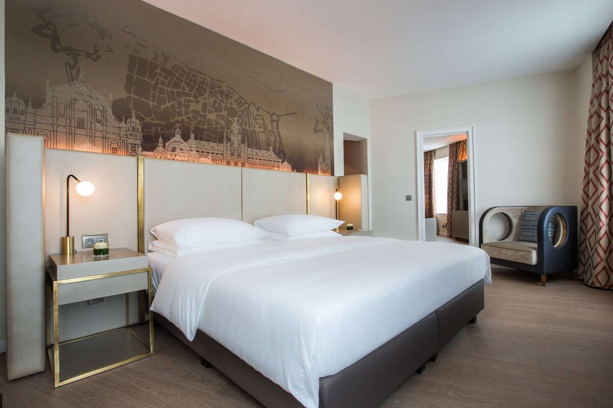 Habitación de hotel con una cama grande, mesitas de noche, un mural en la pared, una zona de estar y una cortina estampada.